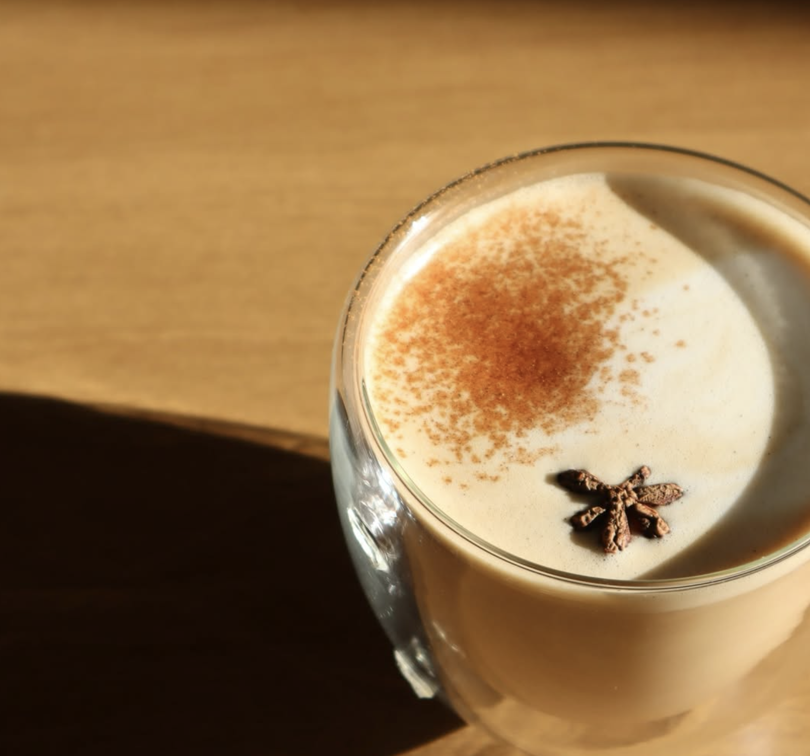 A glass mug of frothy beverage topped with a sprinkle of cinnamon and a star anise pod, on a wooden surface.