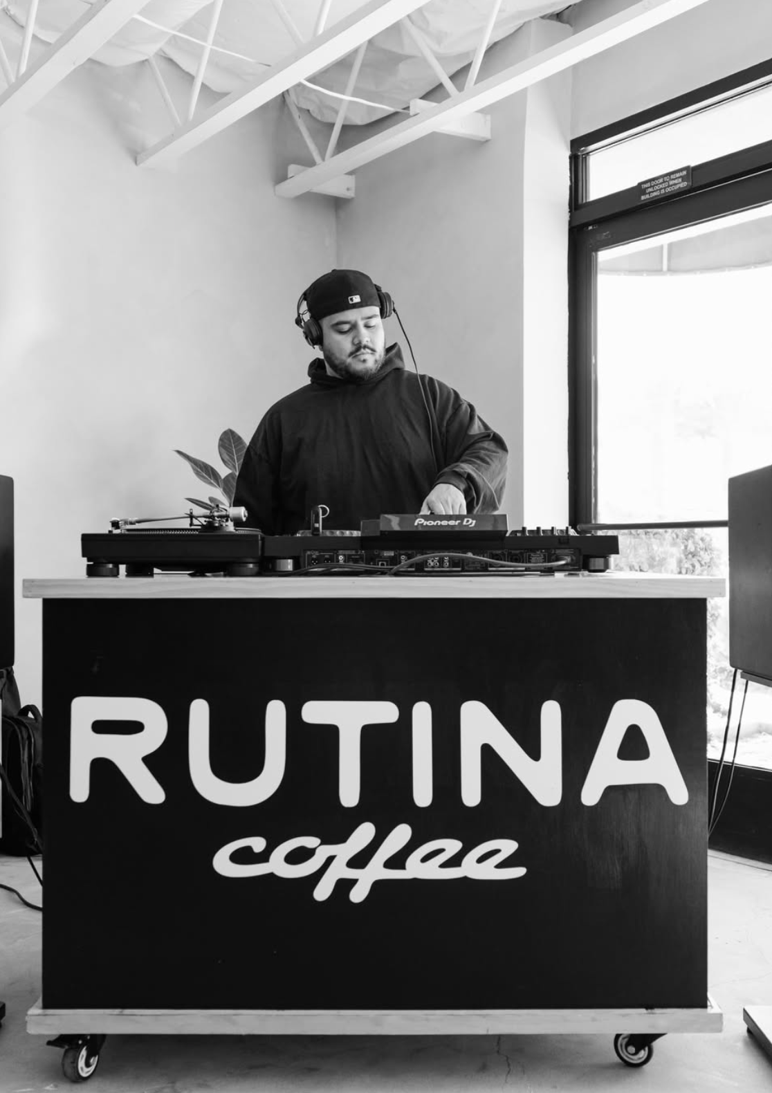 A DJ wearing headphones and a cap, mixing music at a booth with a sign that reads 'RUTINA coffee' inside a modern cafe or studio with large windows.