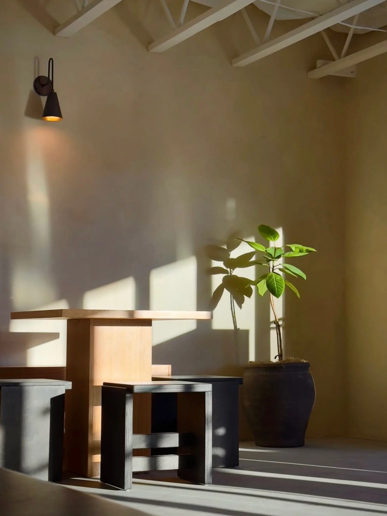 Interior of a room with a wall sconce, a small wooden table, a modern stool, and a potted plant with sunlight casting shadows.