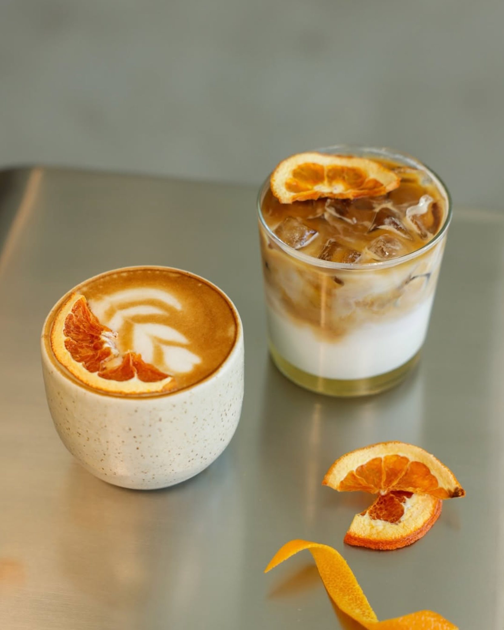 A cup of coffee with latte art and a slice of orange on top, a glass of iced coffee with cream and a slice of orange, and pieces of orange peel on a silver table.