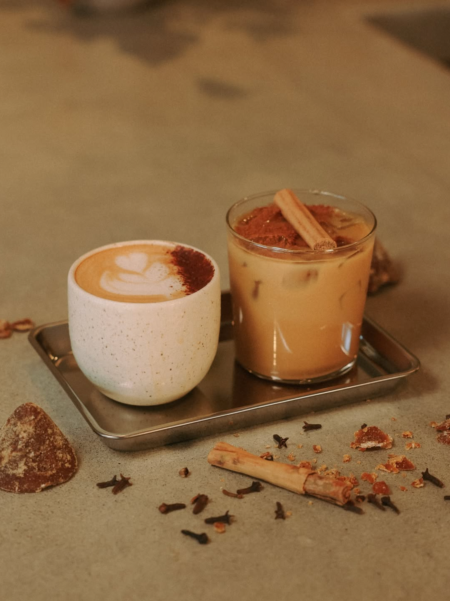A cup of latte art and a glass of iced coffee with a cinnamon stick on a tray, surrounded by scattered cinnamon sticks, cloves, and a piece of salted caramel