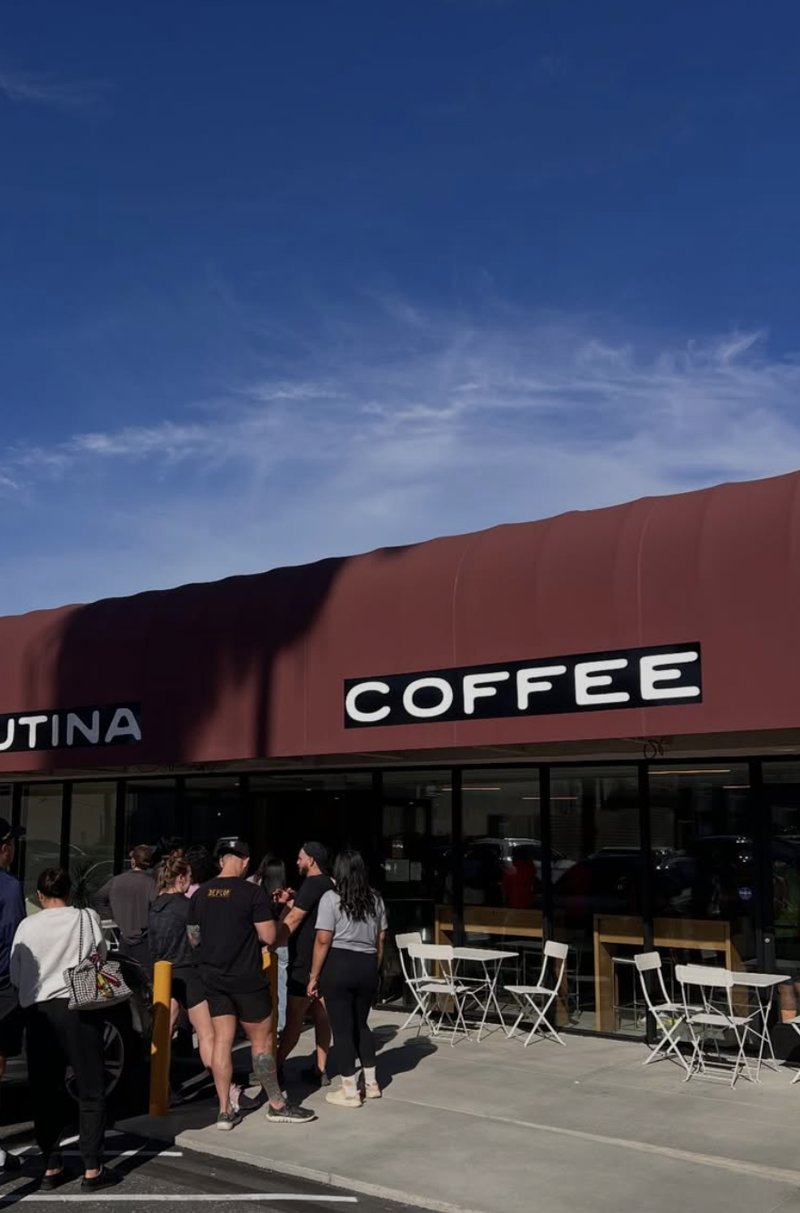 People standing in line outside a coffee shop with a red awning in front of glass windows and outdoor tables, under a partly cloudy sky.