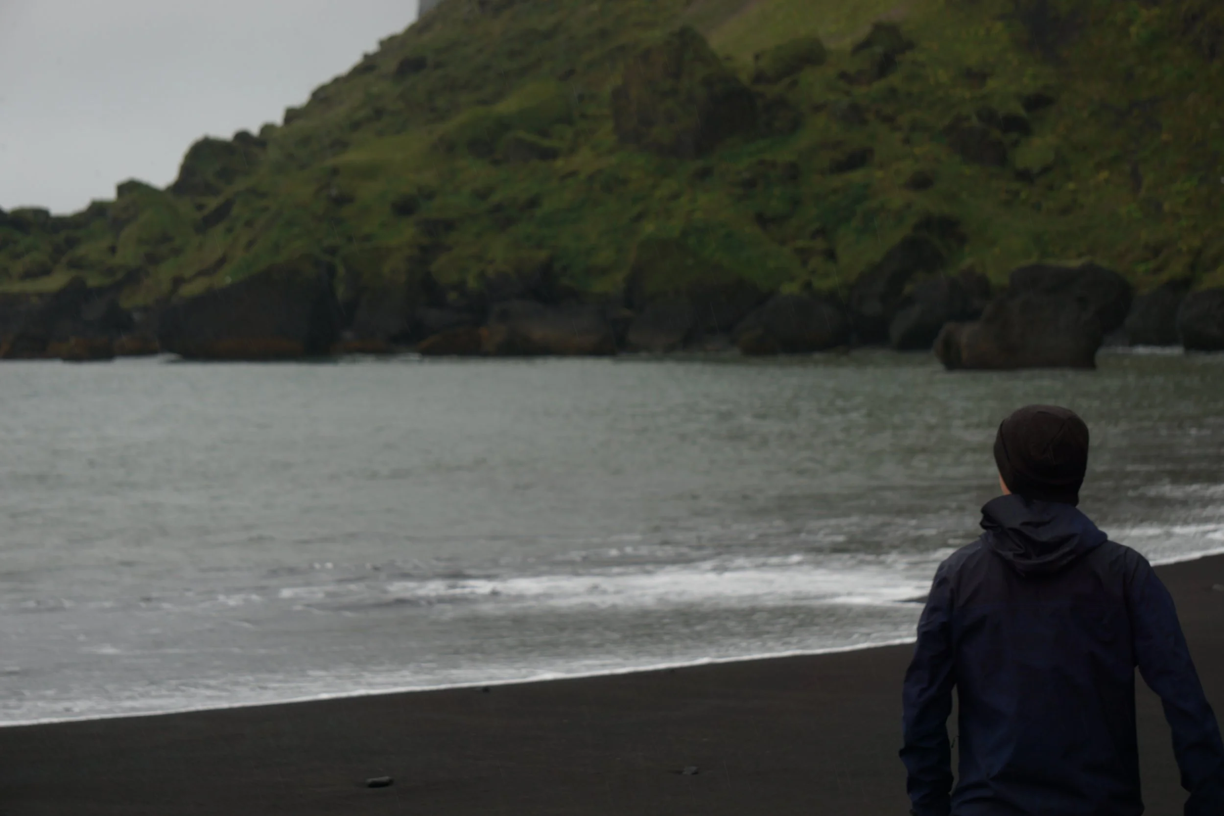 A person wearing a dark jacket and beanie looking at a rocky, green hillside across a body of water on a cloudy day.