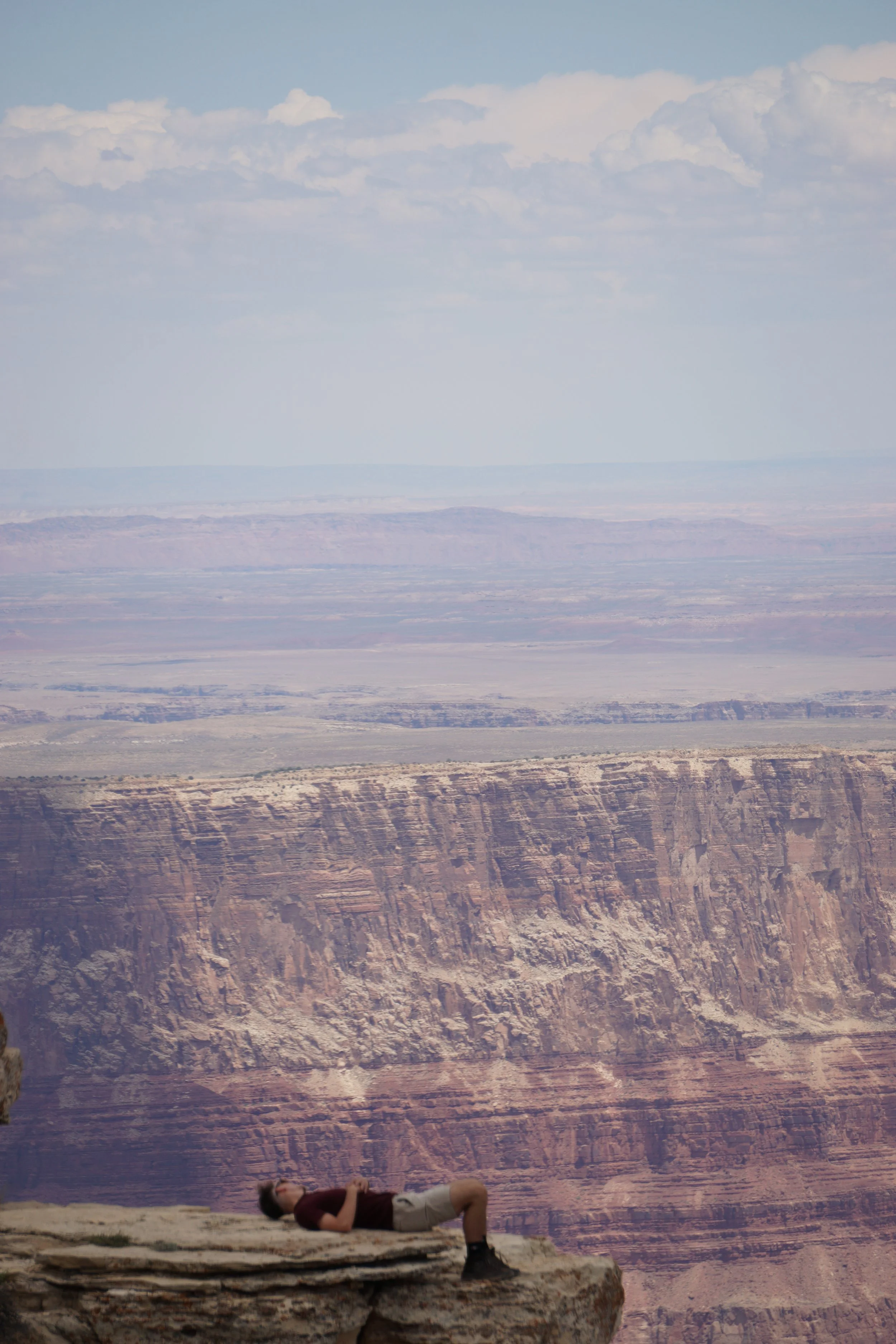 Person lying down on a rock ledge overlooking the Grand Canyon with layered rock formations and a vast sky in the background.