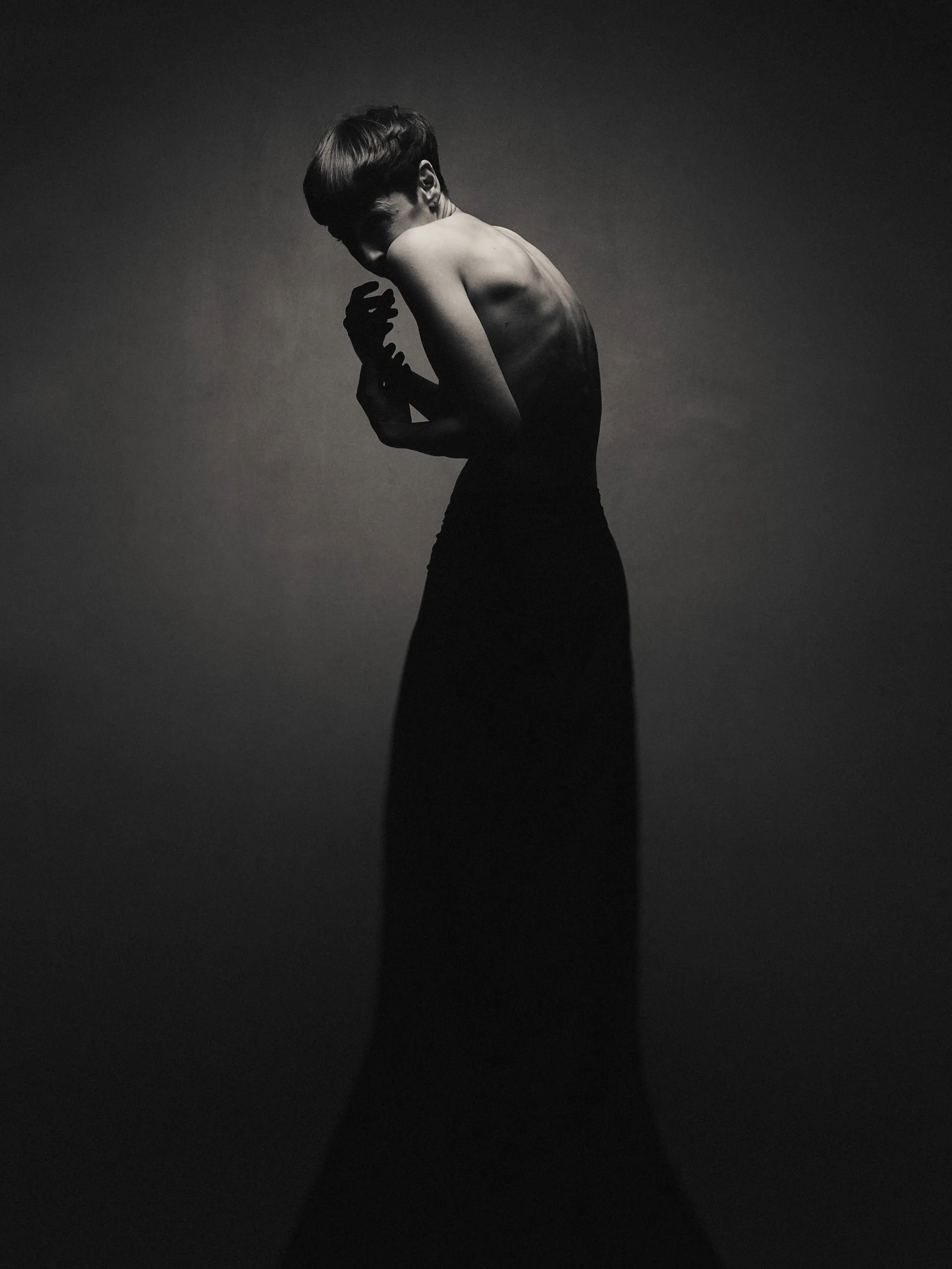Photo en noir et blanc d'une femme nue avec un haut de robe ou jupe longue, qui se tient de côté, la tête baissée et les mains près du visage, dans une pose artistique dramatique.
