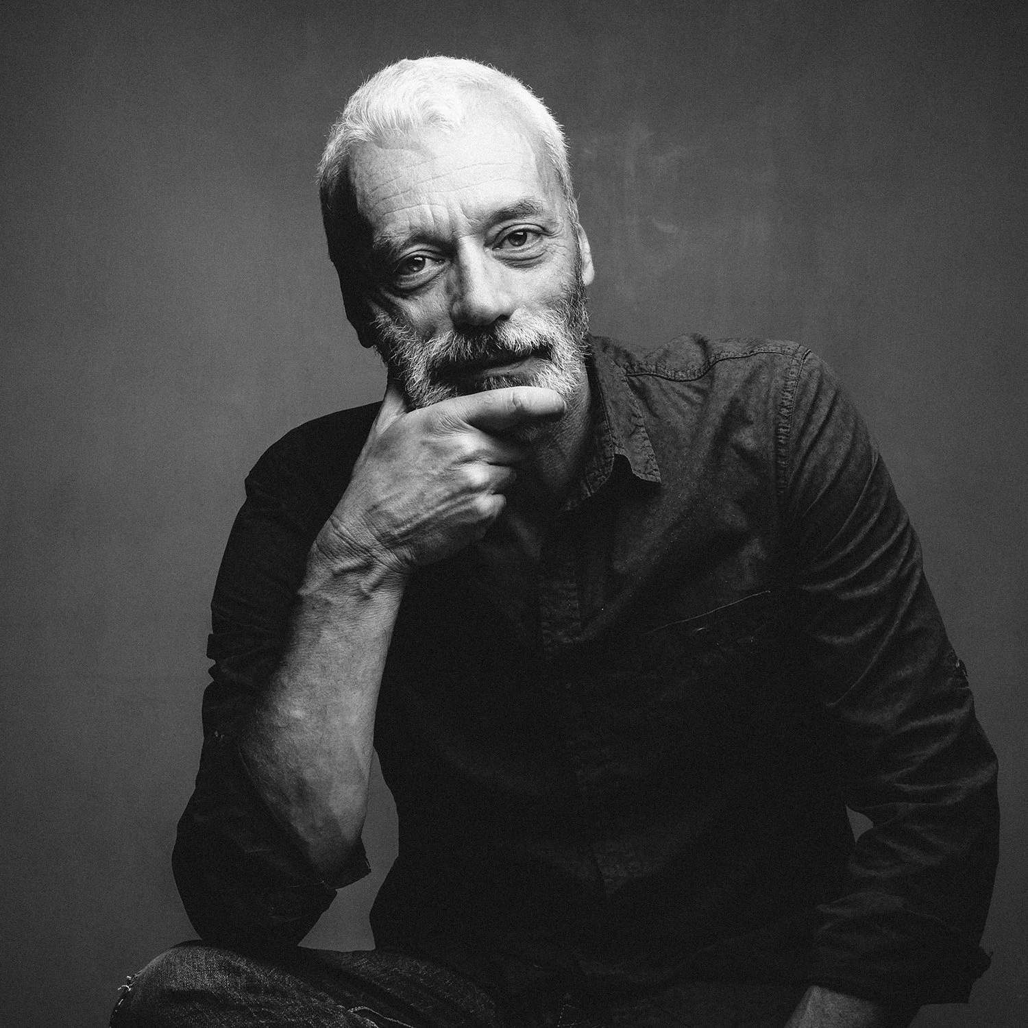 Portrait d'un homme âgé avec une barbe blanche, portant une chemise noire, regardant pensivement l(appareil photo, dans une image en noir et blanc.