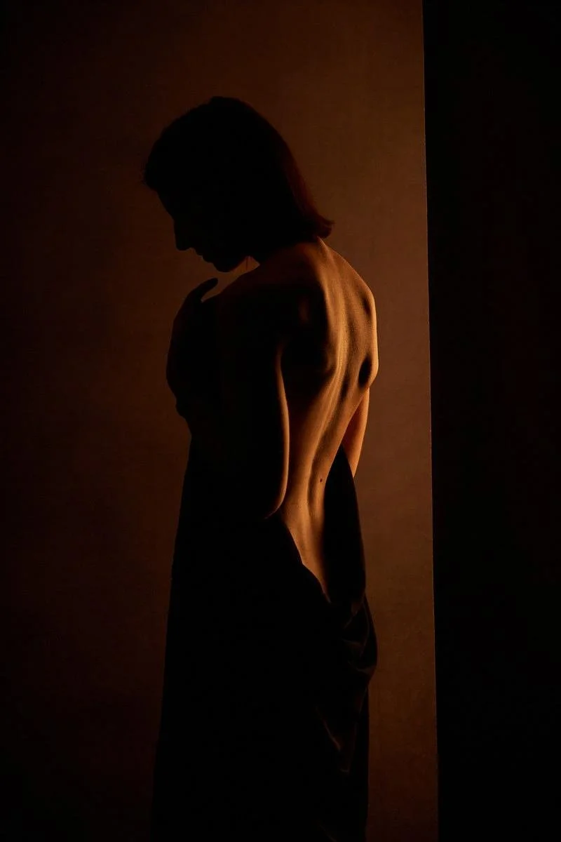 Silhouette d'une femme nue de profil et de dos dans un éclairage tamisé, créant une ambiance mystérieuse.