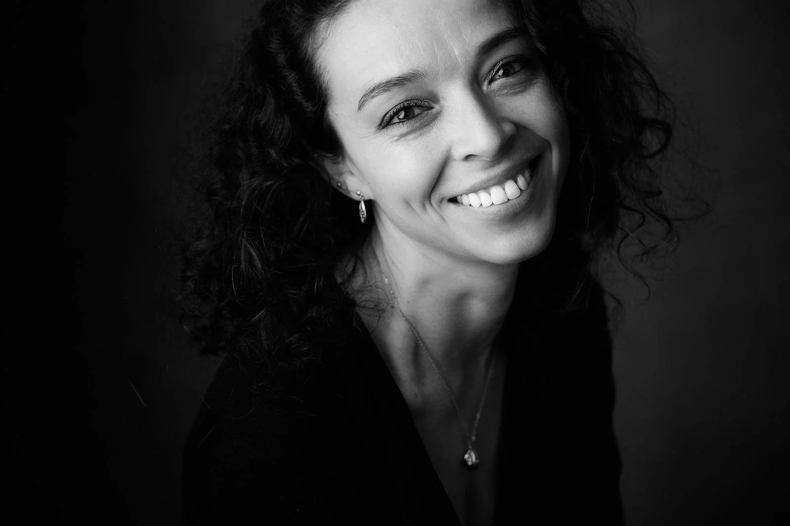 Portrait en Noir et Blanc d'une femme souriante avec des cheveux bouclés, portant des boucles d'oreilles et un collier.