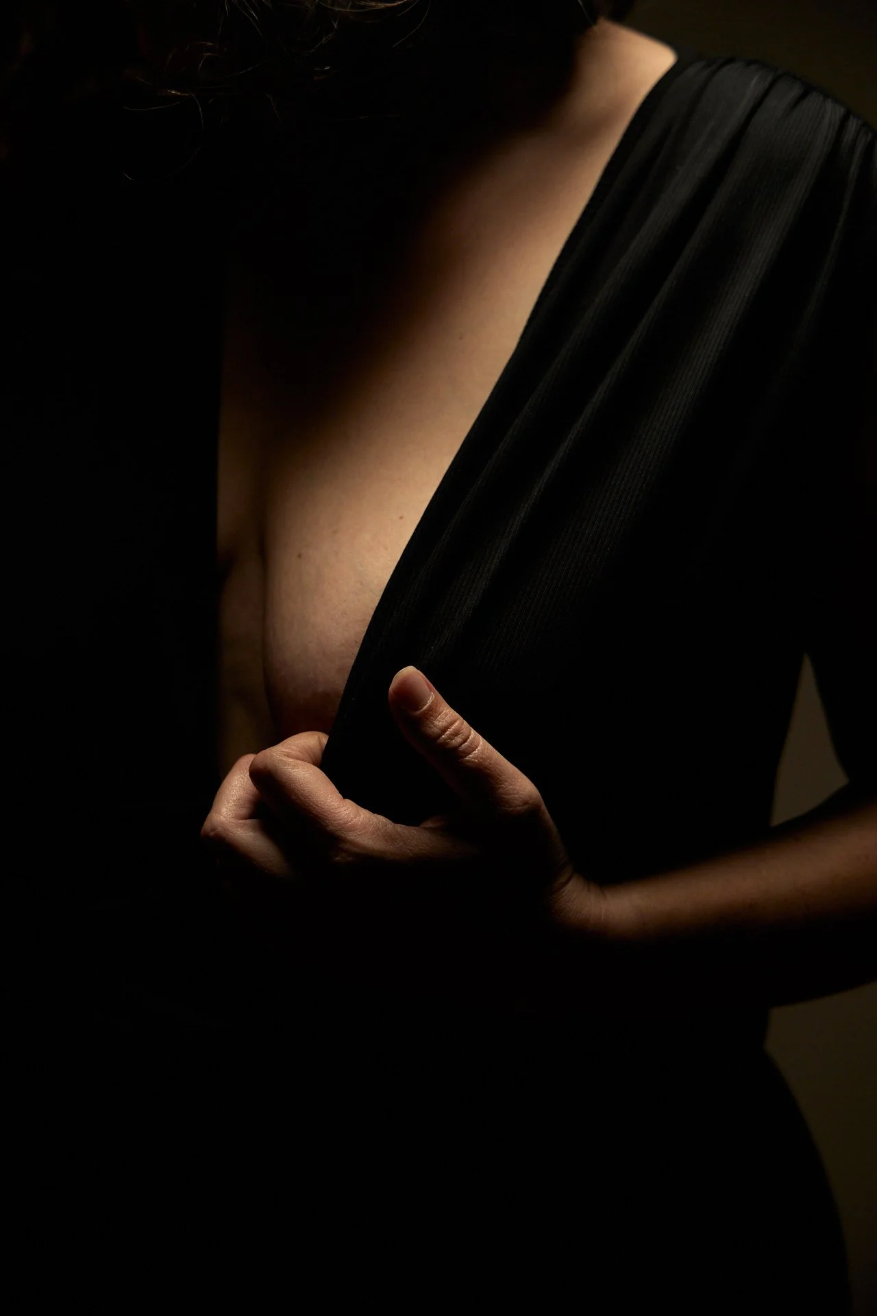 Une personne en noir dont la main tient un vêtement en tissu noir à rayures, en révélant partiellement une peau nue. L'éclairage est sombre, avec une mise en valeur de la peau et du tissu.