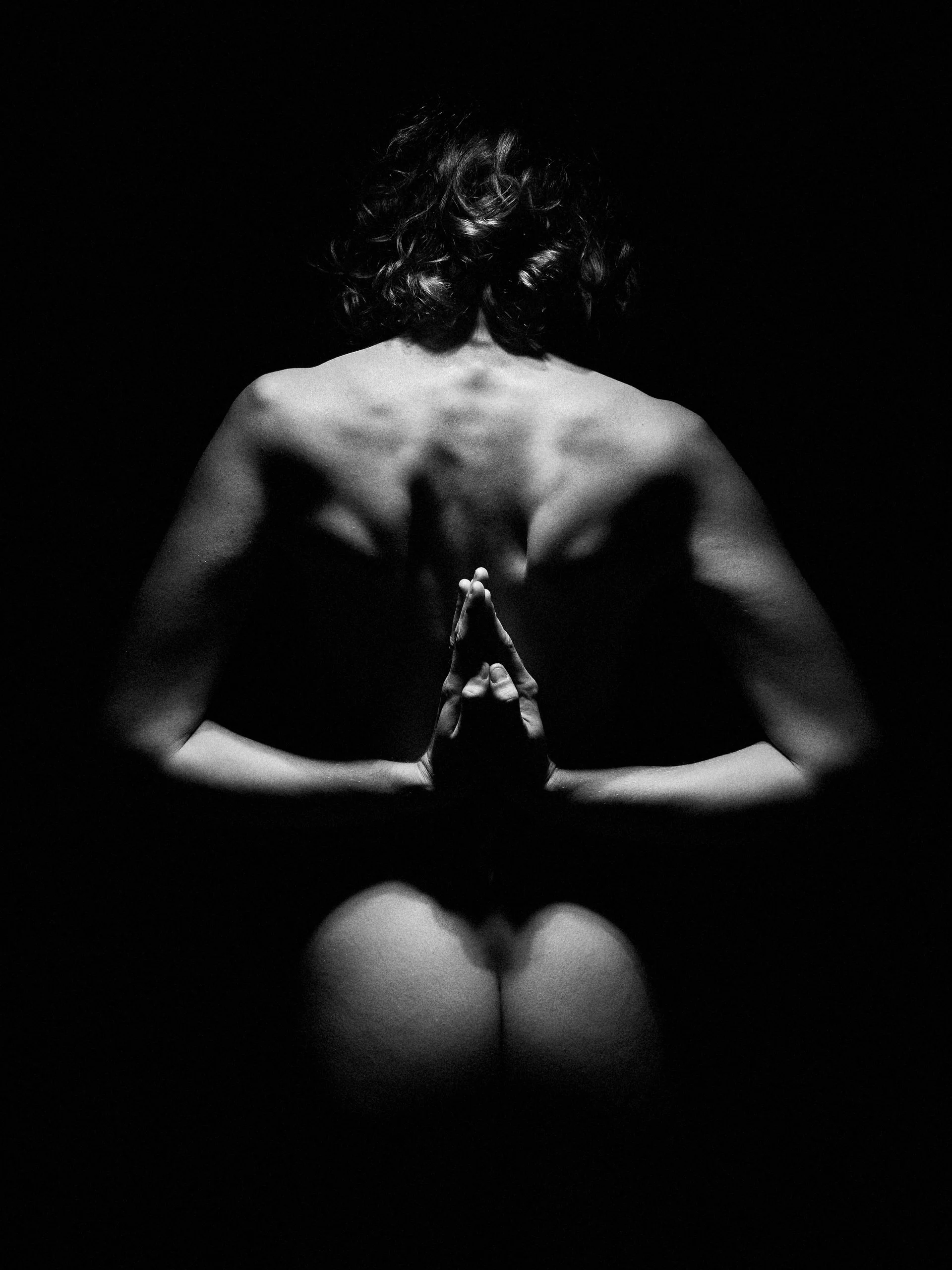 Une personne nue vue de dos avec les bras levés en position de prière, laissant apparaître ses épaules et son dos, dans un contexte sombre et en noir et blanc.