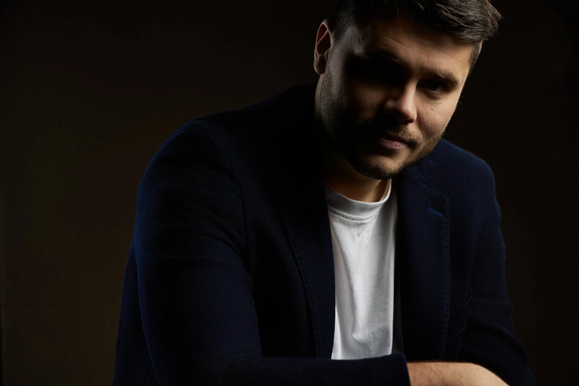 Jeune homme avec des cheveux courts, portant une veste noire et un t-shirt blanc, posé contre un fond sombre.