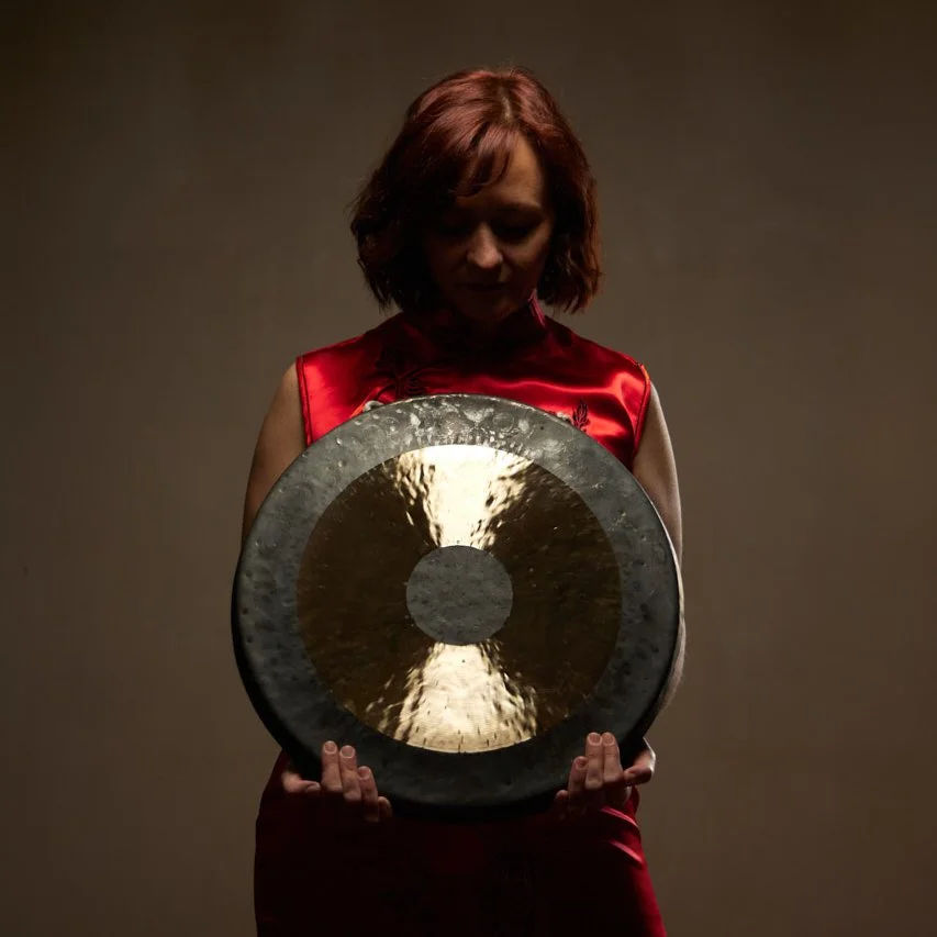 Une femme aux cheveux roux portant une robe rouge, tenant un gong en métal brillant dans ses mains, avec un fond sombre.