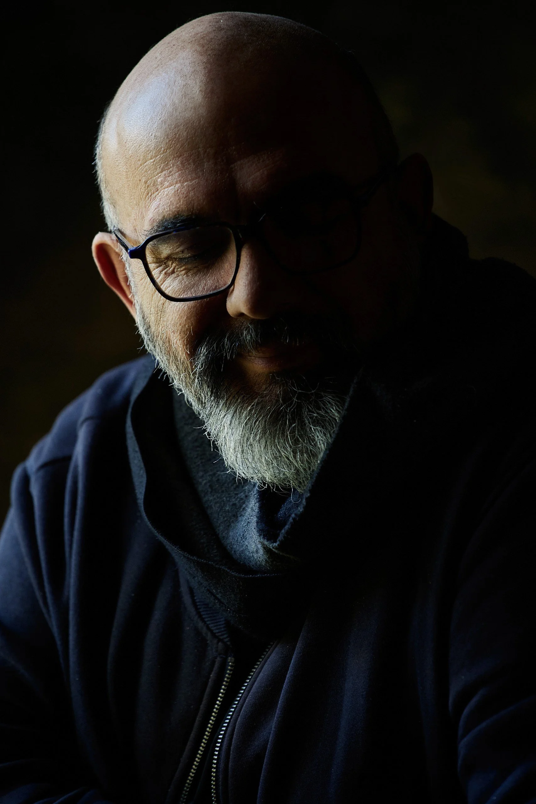 Un homme barbu avec lunettes, dans une lumière tamisée, portant un sweat à capuche noir.