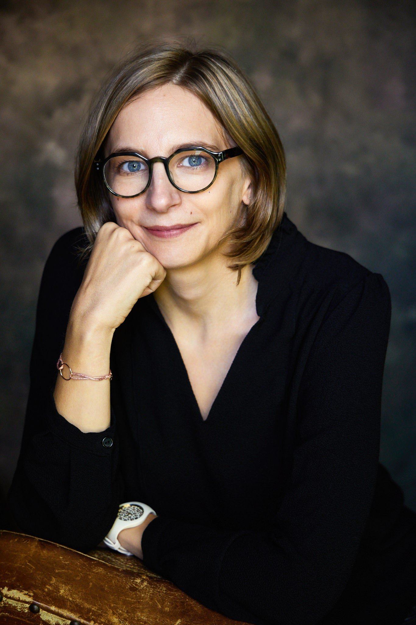 Une femme avec des lunettes rondes, portant un chemisier noir, posant avec la main sous le visage, sourire, dans un fond sombre.