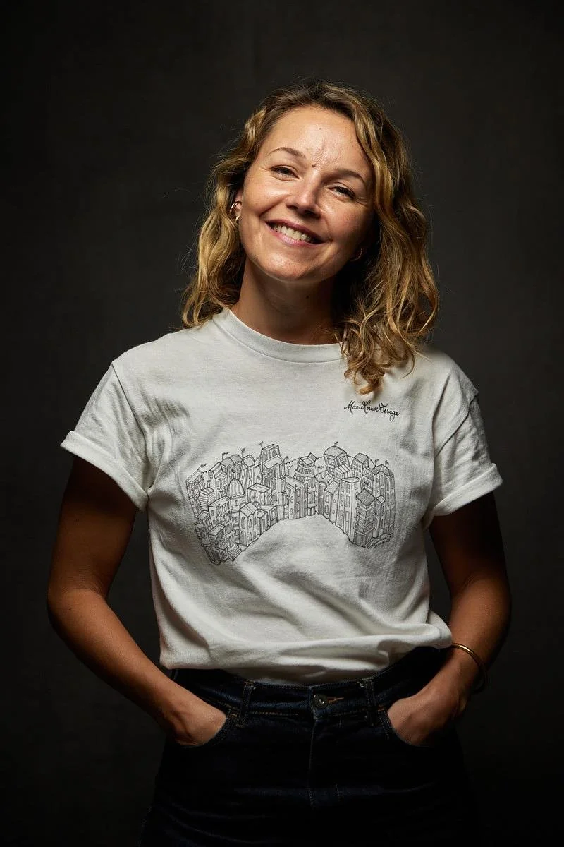Jeune femme souriante portant un t-shirt blanc avec une illustration de bâtiments de ville dessus, posée contre un fond noir.