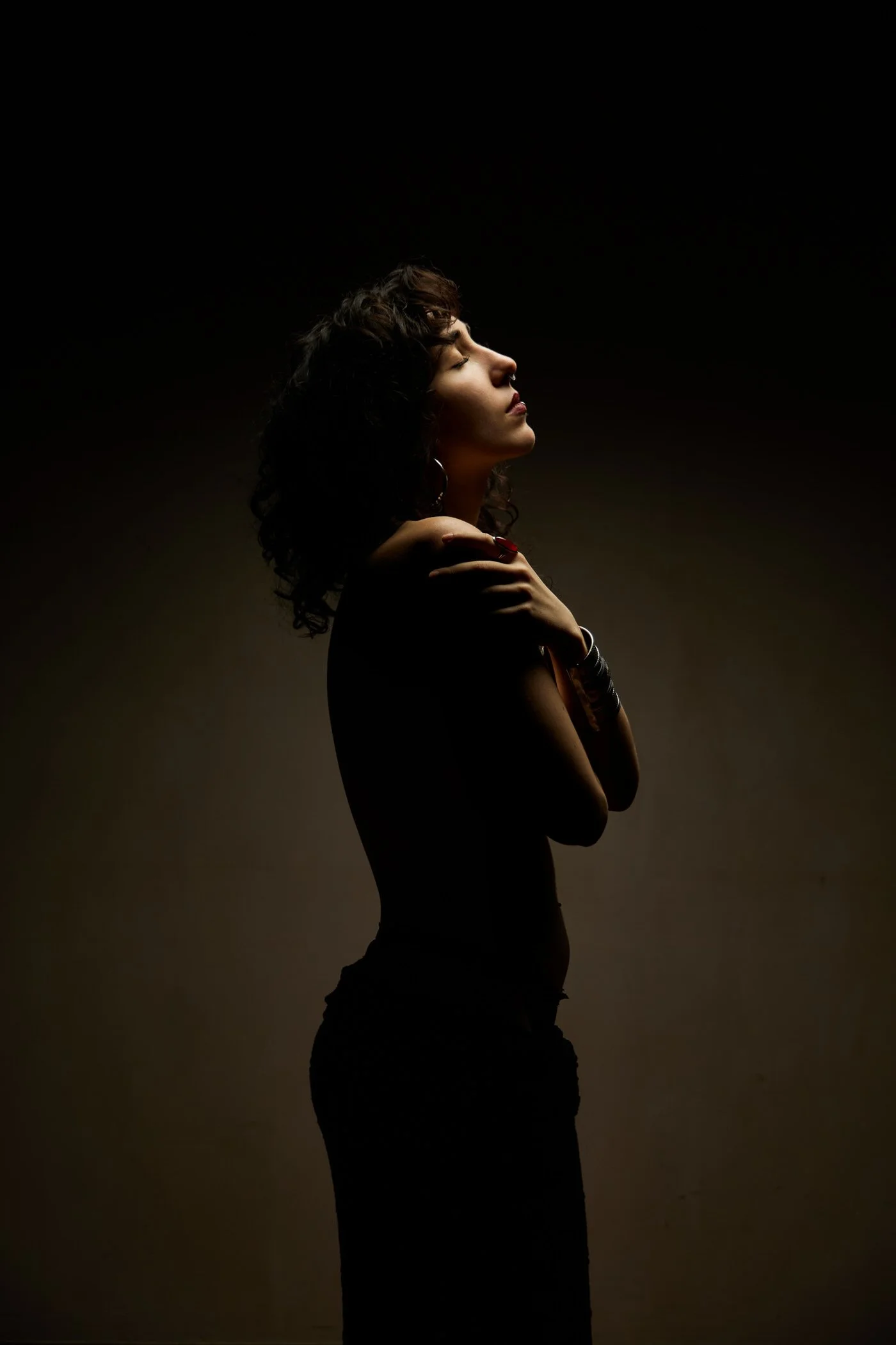 Profil d'une femme en posture élégante, bras croisés, avec des cheveux bouclés, sous un éclairage tamisé, fond sombre.