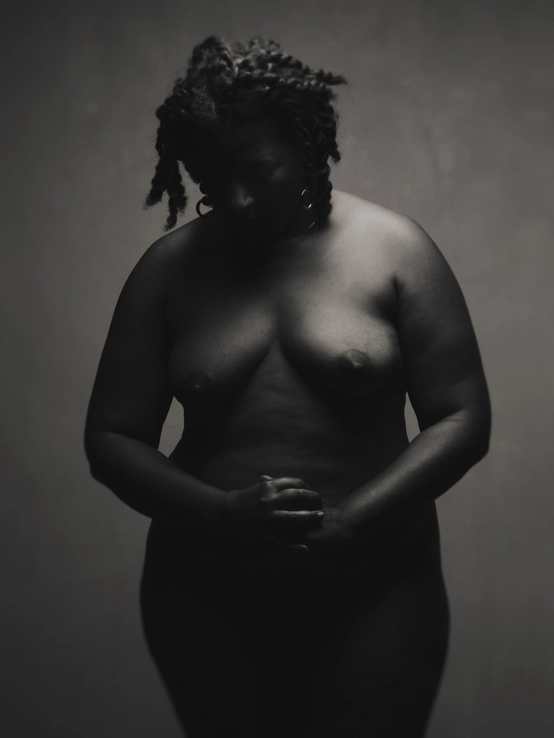Une femme nue, de dos, avec des cheveux dreadlocks, se tient la tête baissée, en position de prière ou de réflexion, dans une photographie en noir et blanc.