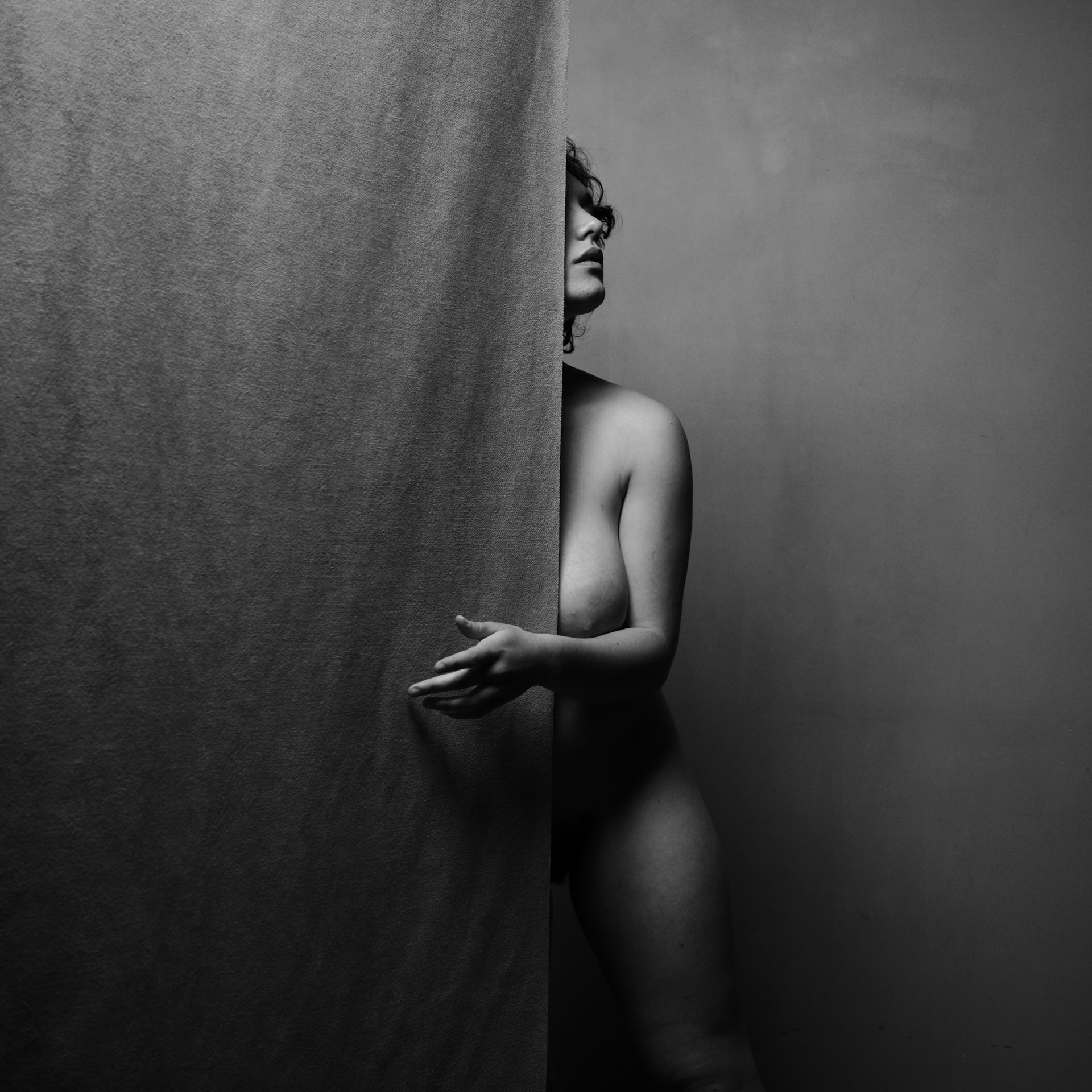 Une femme nue en noir et blanc, partiellement cachée derrière un rideau, avec un fond gris