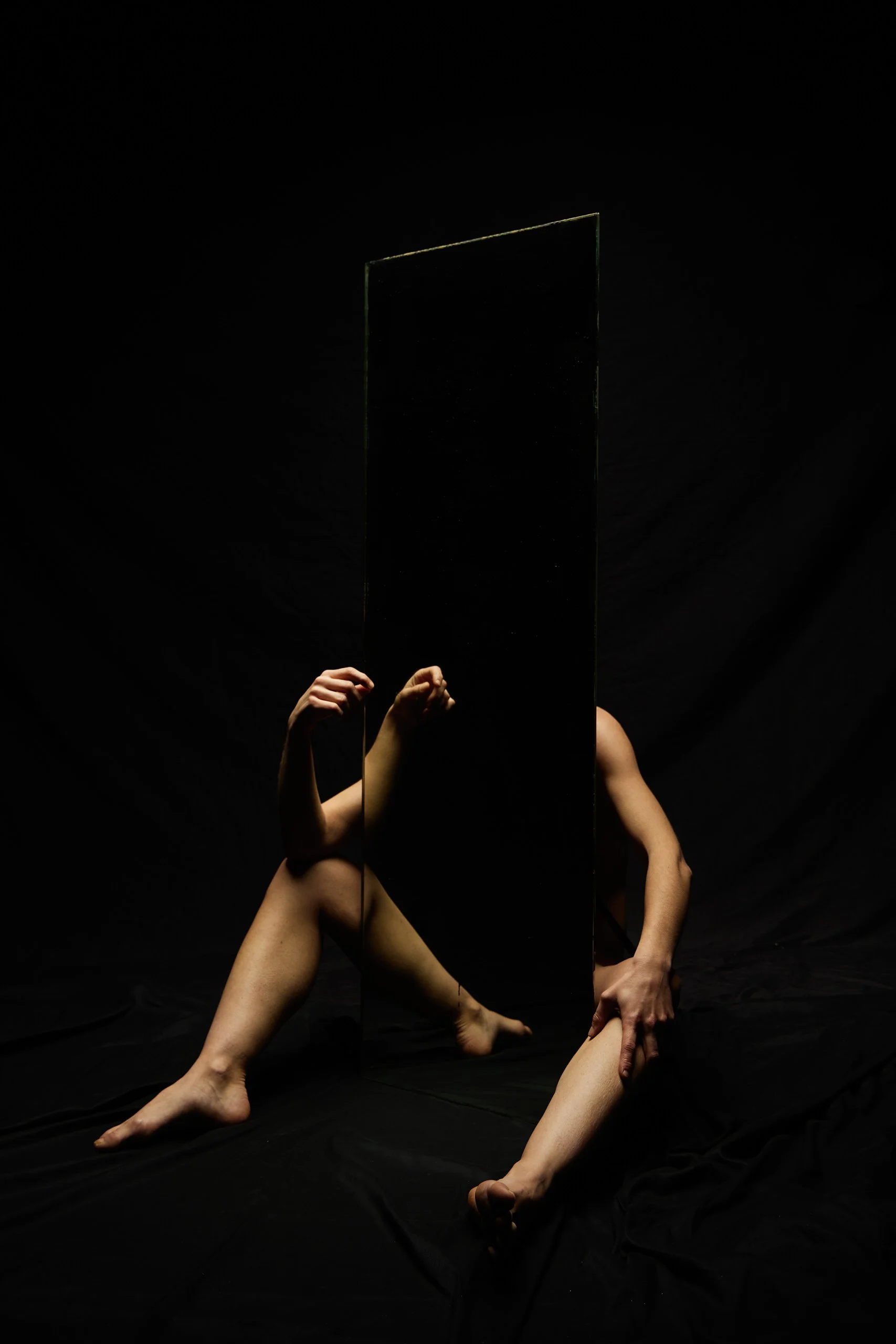 Une personne nue assise sur le sol noir, tenant un miroir rectangulaire vertical qui couvre une partie de son corps. Le fond est noir, créant un contraste avec la peau de la personne.