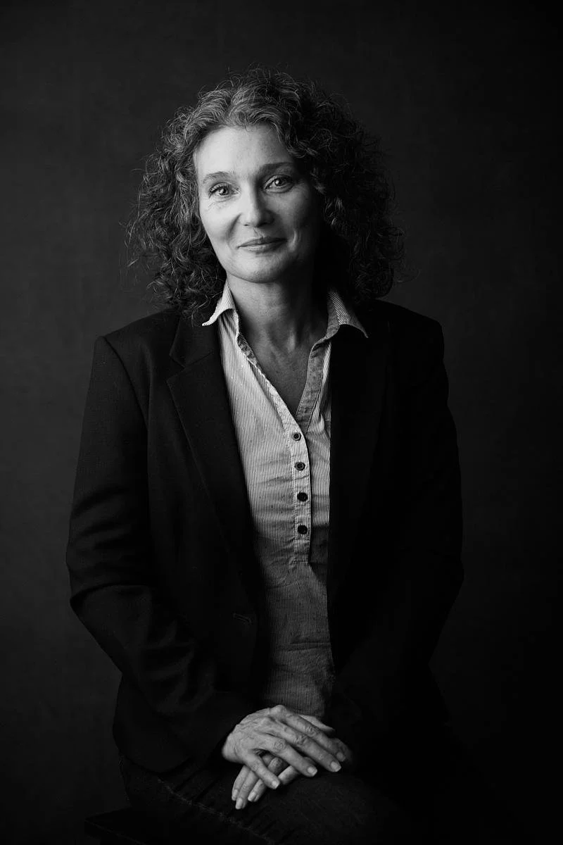 Portrait en noir et blanc d'une femme avec des cheveux bouclés, portant une veste noire et une chemise rayée, assise avec les mains croisées.