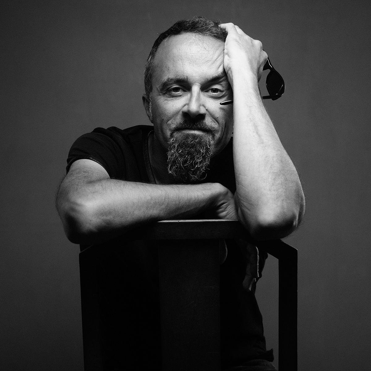 Homme souriant avec barbe, tenant des lunettes de soleil, posé sur une table, en noir et blanc.