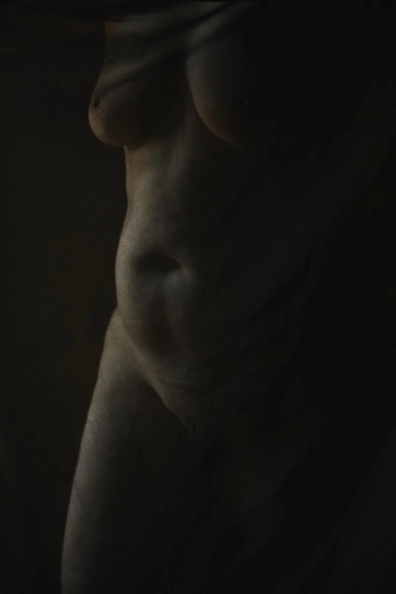 Vue partielle d'un corps de femme nu derrière un voile, en basse lumière.