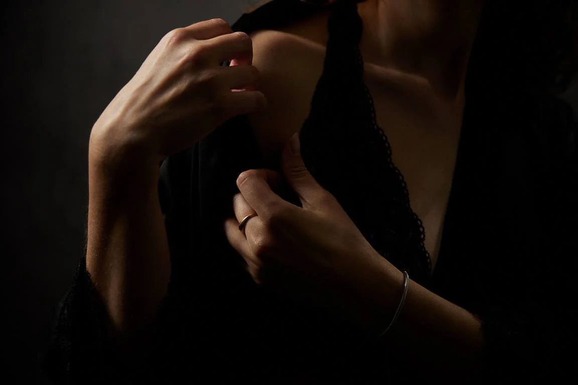 Close-up de deux mains féminines, une touchant l'épaule et l'autre tenant la robe noire en dentelle, sur fond noir.