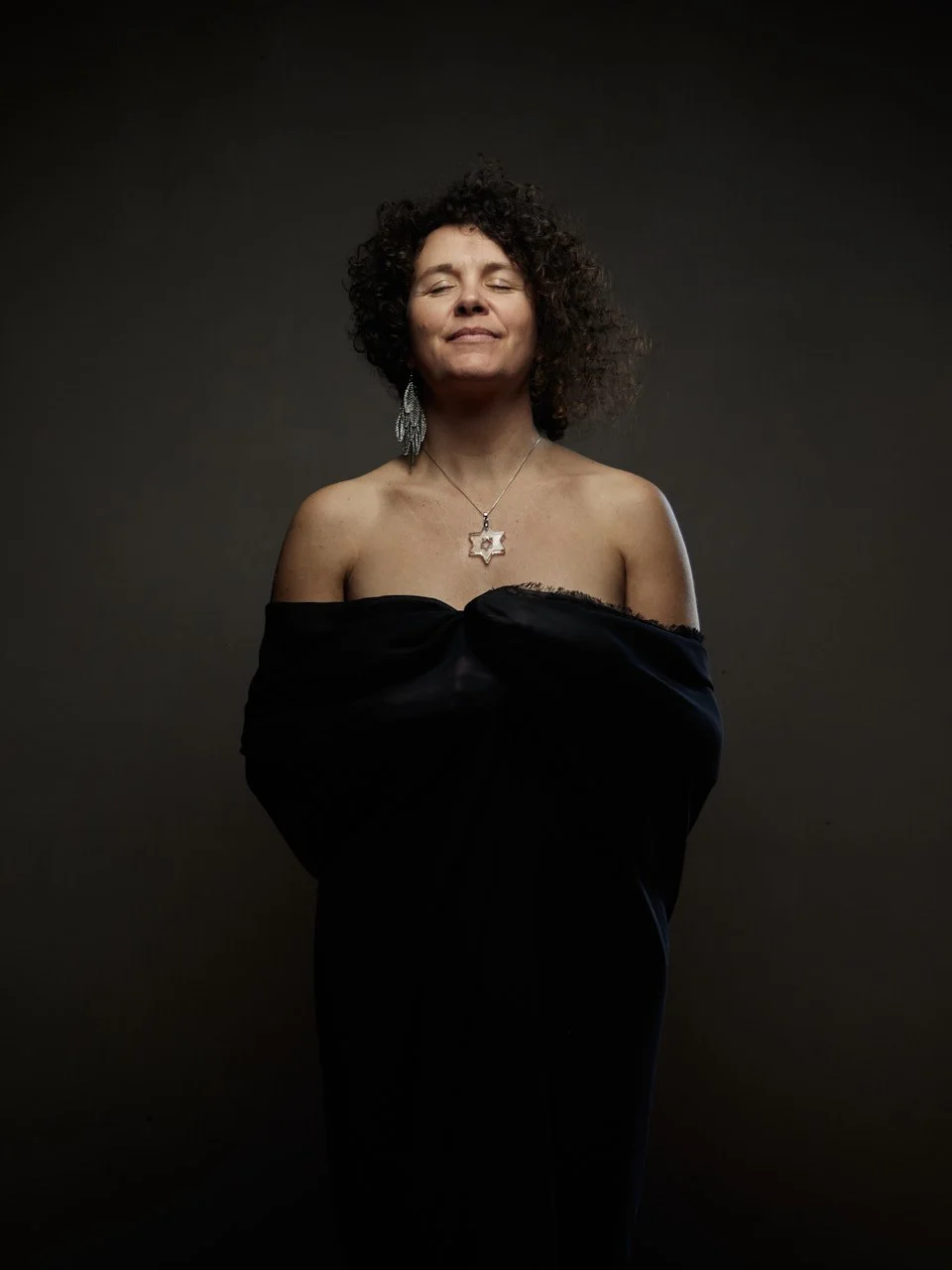 Femme souriante, yeux fermés, portant une robe noire avec des bijoux, debout contre un fond sombre.