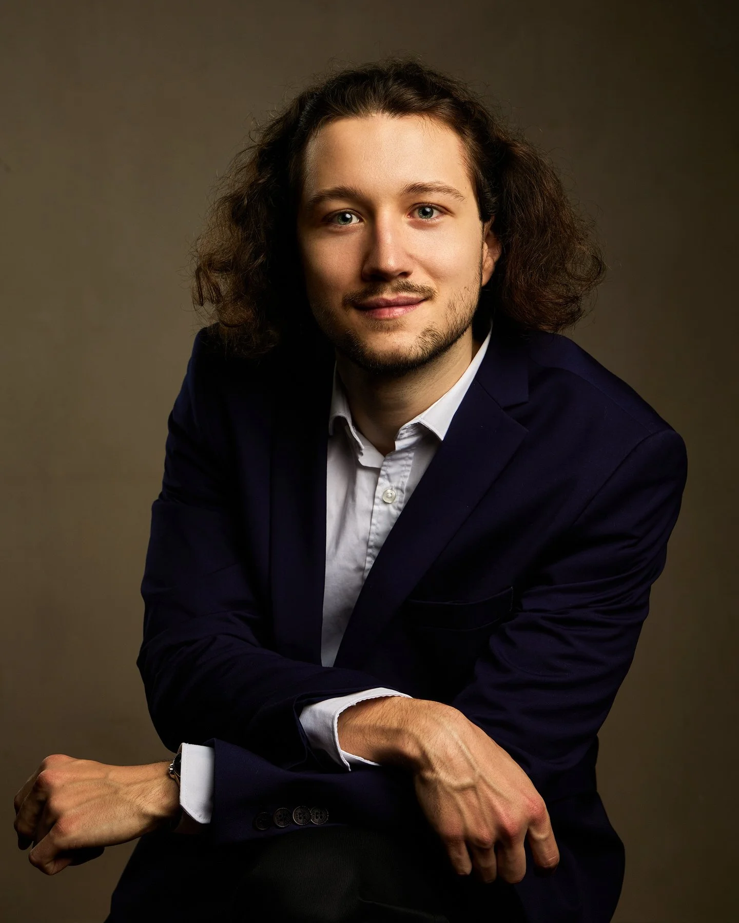 Portrait d'un homme avec des cheveux longs et bouclés, portant un blazer bleu foncé et une chemise blanche, assis contre un fond neutre.