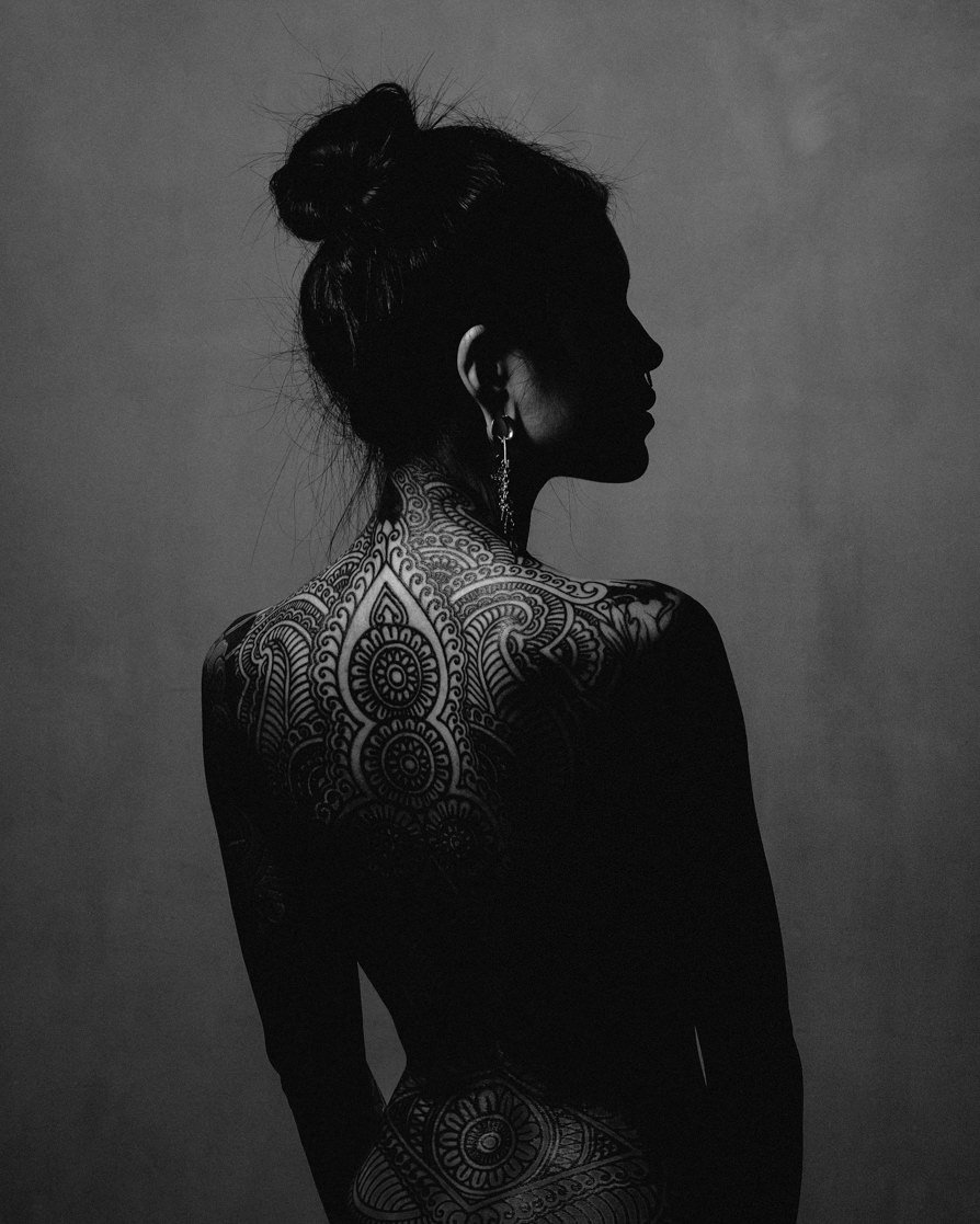 Silhouette noire et blanche d'une femme vue de profil, avec un tatouage complexe sur le dos et le cou, portant une boucle d'oreille longue.