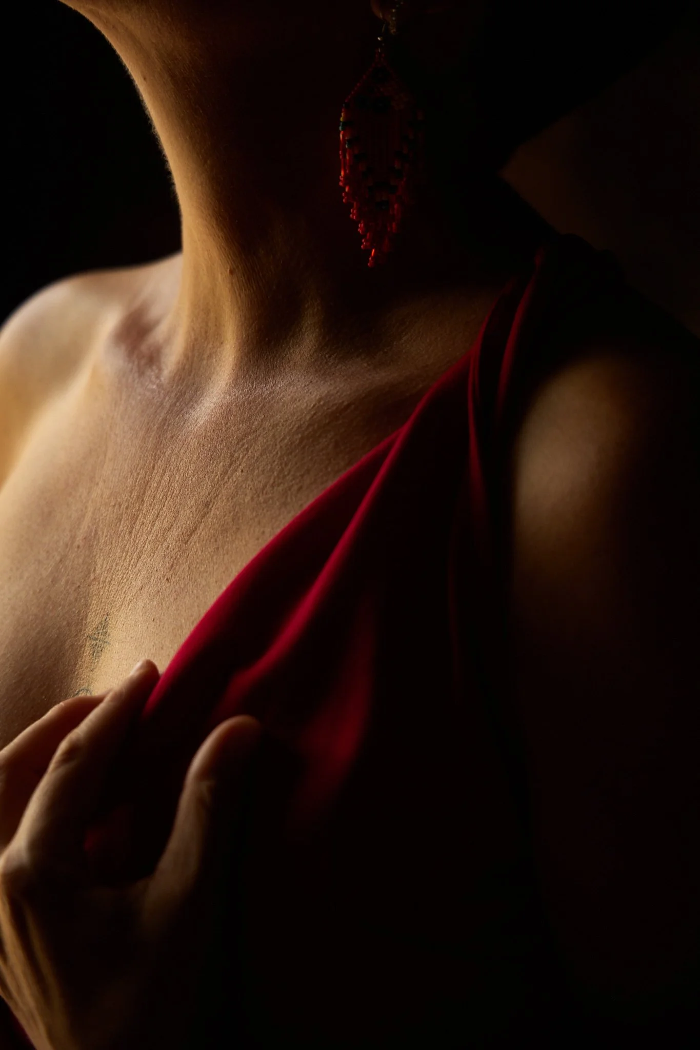 Une personne portant un foulard rouge et une boucles d'oreilles de perles rouges, le visage caché dans l'ombre, mettant en valeur le cou et la main.
