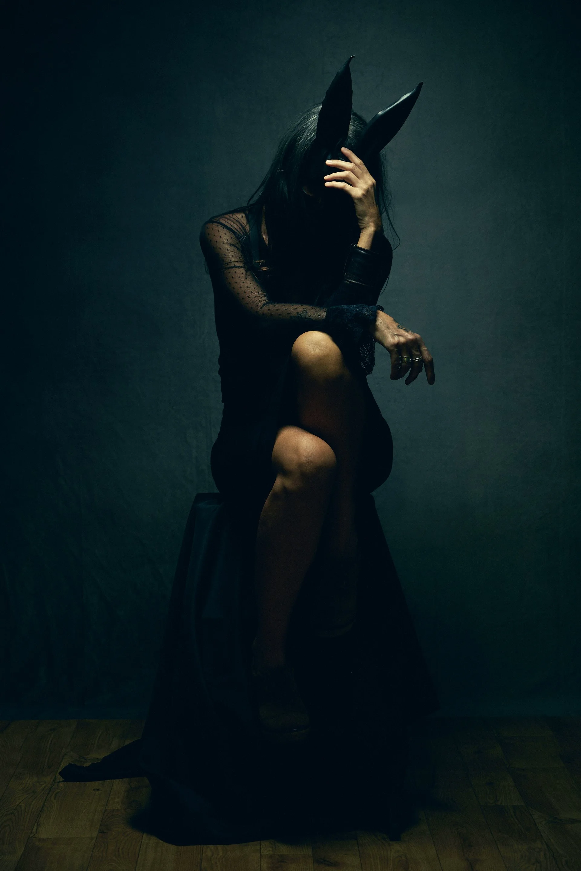 Femme portant des oreilles de lapin en plastique noir, vêtue d'une robe noire transparente avec des manches en dentelle, assise sur un tabouret dans une pièce sombre avec un fond gris, avec une main sur le visage.