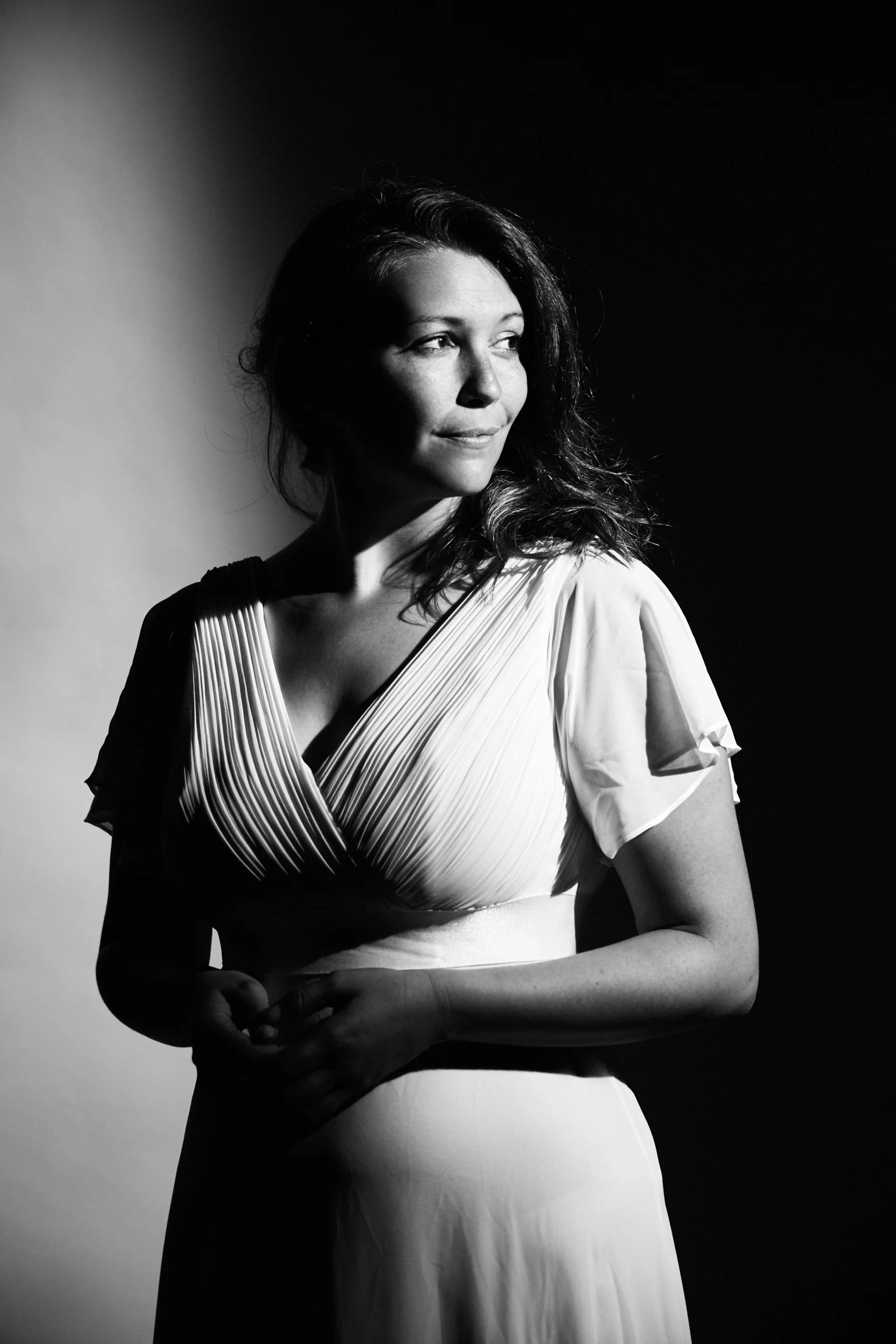 Une femme en robe claire, debout devant un fond en deux tons, avec un éclairage contrasté créant des ombres fortes sur son visage et son corps.