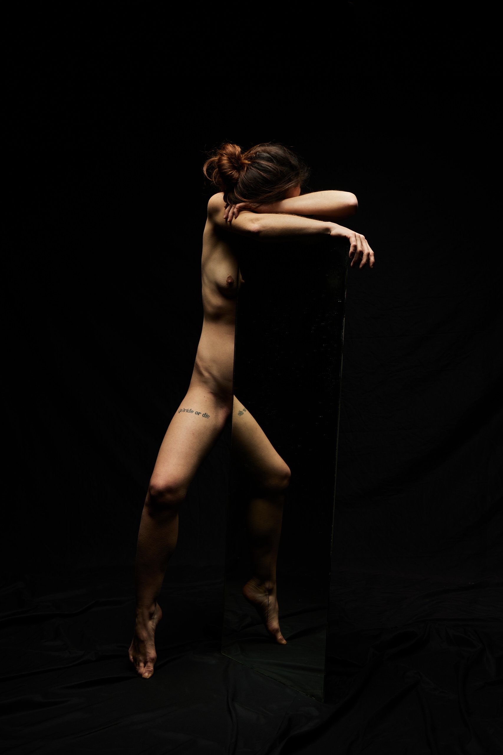 Une femme nue avec des tatouages pose devant un miroir noir dans un fond noir, sa tête repose sur ses bras croisés sur le miroir.