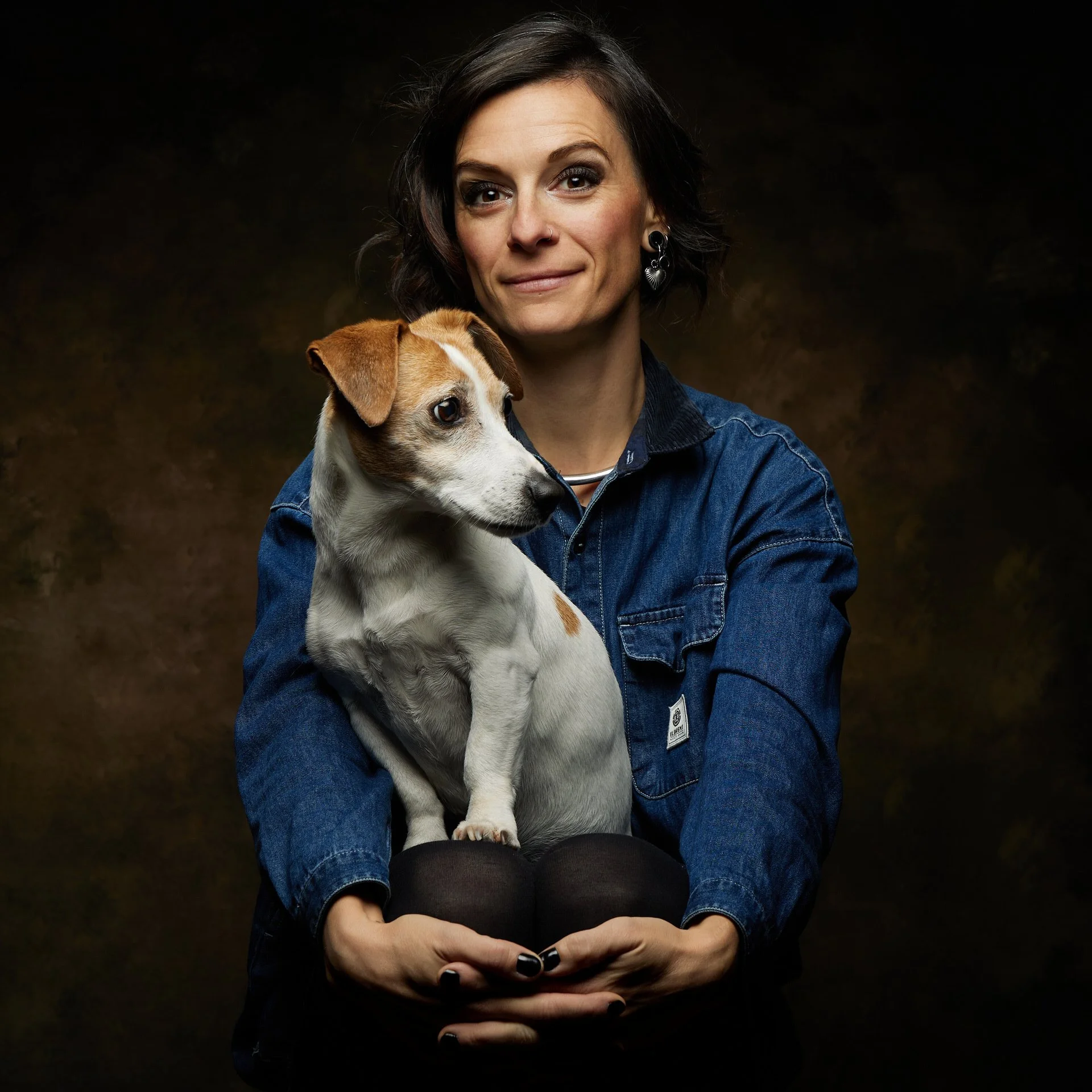 Une femme avec un chien blanc et marron sur ses genoux, portant une veste en jean, en portrait contre un fond sombre.