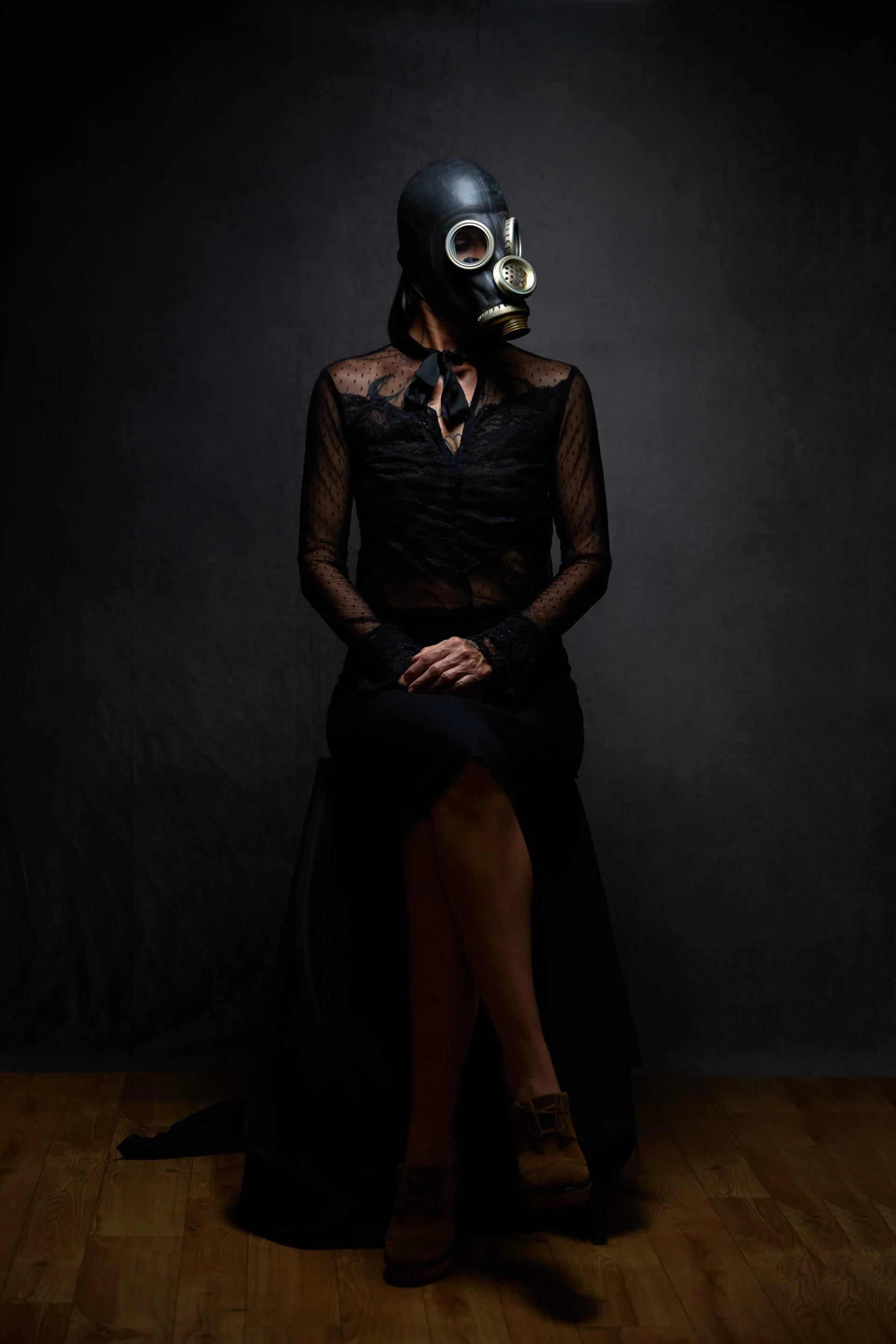 Une personne assise sur une chaise portant un masque à gaz noir et une robe noire en dentelle, avec un fond sombre et un sol en bois.