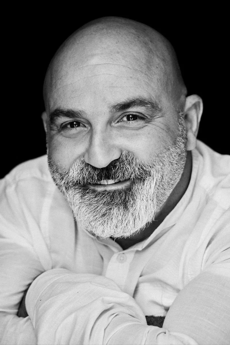 Portrait en noir et blanc d'un homme souriant avec une barbe grise, portant une chemise blanche, regardant vers l'objectif.