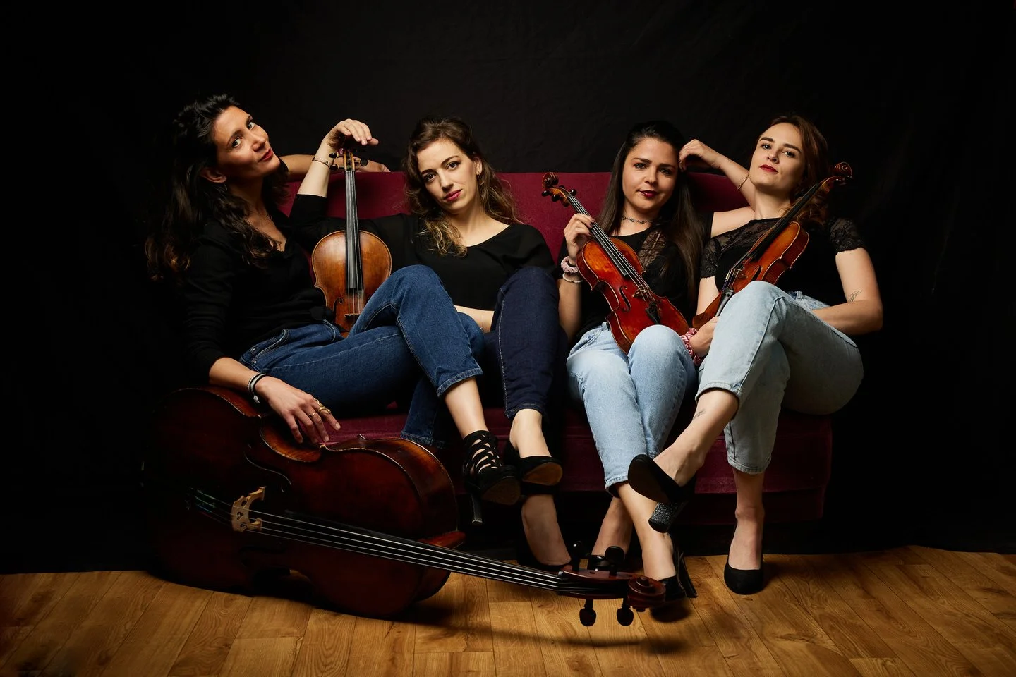 Quatre femmes assises sur un canapé rouge, tenant ou entourant des violons, avec un violoncelle posé au sol devant elles. L'arrière-plan est noir et le sol en bois.