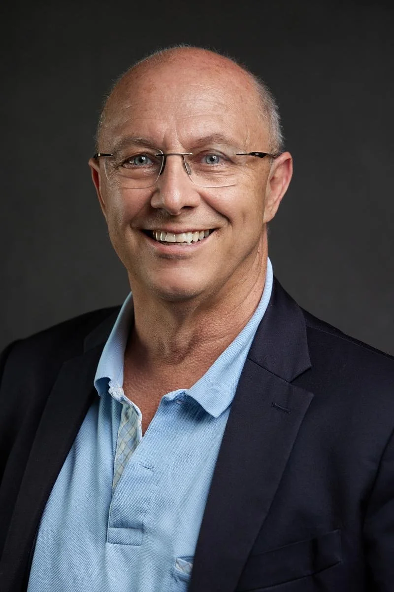 Un homme souriant portant des lunettes, un costume et une chemise bleue, fond gris foncé.