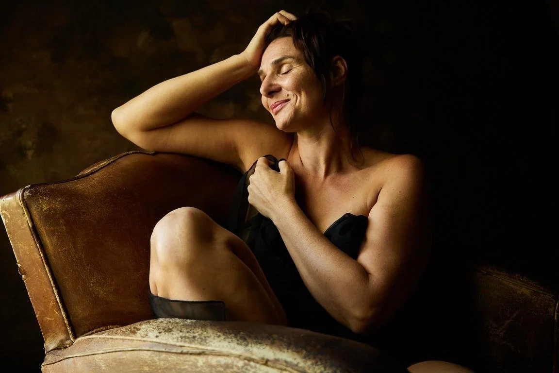 Une femme souriante assise sur un vieux canapé en cuir, avec la main sur la tête et tenant un vêtement noir contre sa poitrine, dans une ambiance chaleureuse et intime.