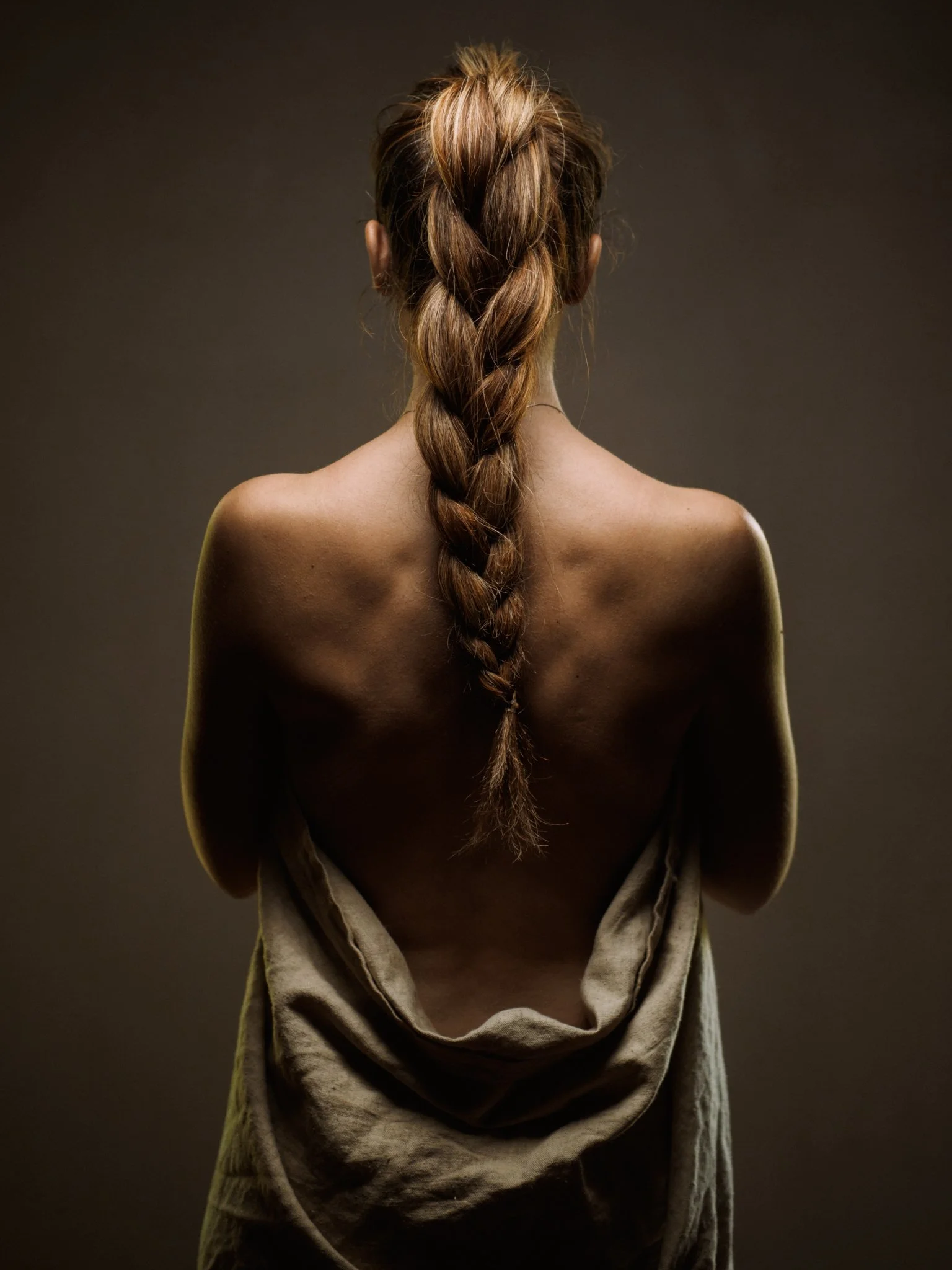 Vue arrière d'une femme nue avec une longue natte brune, tenant un tissu beige autour de ses hanches, contre un fond sombre.