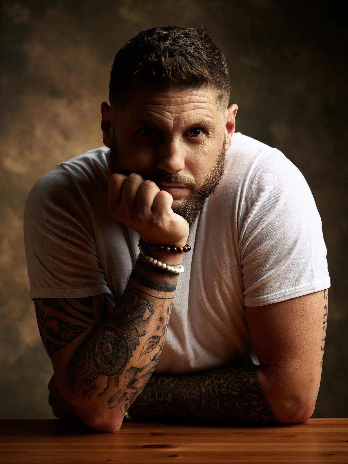 Un homme avec une barbe et tatouages, portant un t-shirt blanc, regarde fixement la caméra, posant avec son bras sur une table en bois, dans une lumière douce et un fond sombre.