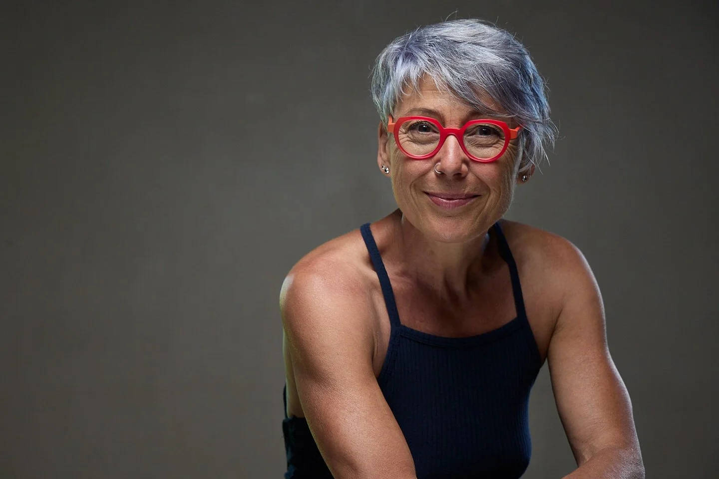 Une femme âgée souriante avec des cheveux gris courts, portant des lunettes rouges et un haut noir à fines bretelles, posé contre un fond gris.