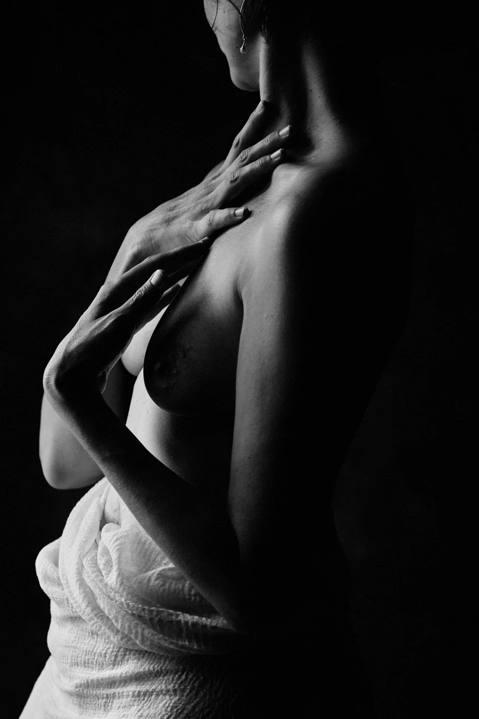 Une femme nue en noir et blanc, touchant son cou et son épaule, avec un fond sombre.