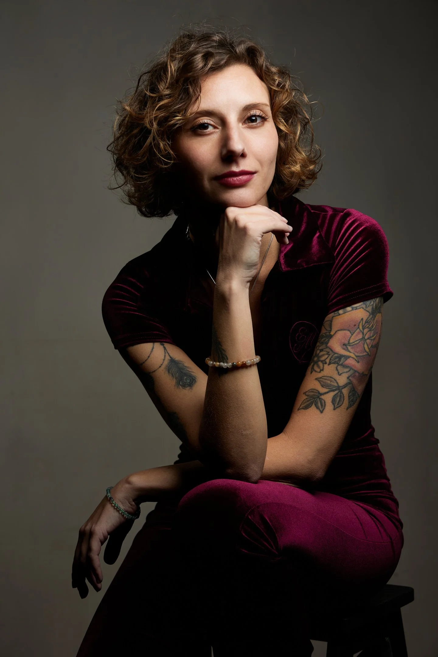 Une femme aux cheveux bouclés bruns, portant une vêtement en velours pourpre, posant avec sa main sous le menton, avec des tatouages sur les bras, un bracelet en perles, et une bague, contre un fond neutre.