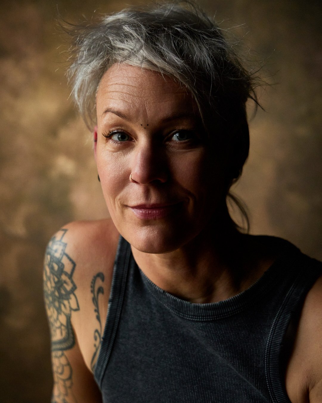 Portrait d'une femme avec des cheveux clairs, un tatouage sur l'épaule, portant un débardeur noir, souriant légèrement, devant un fond flou. 