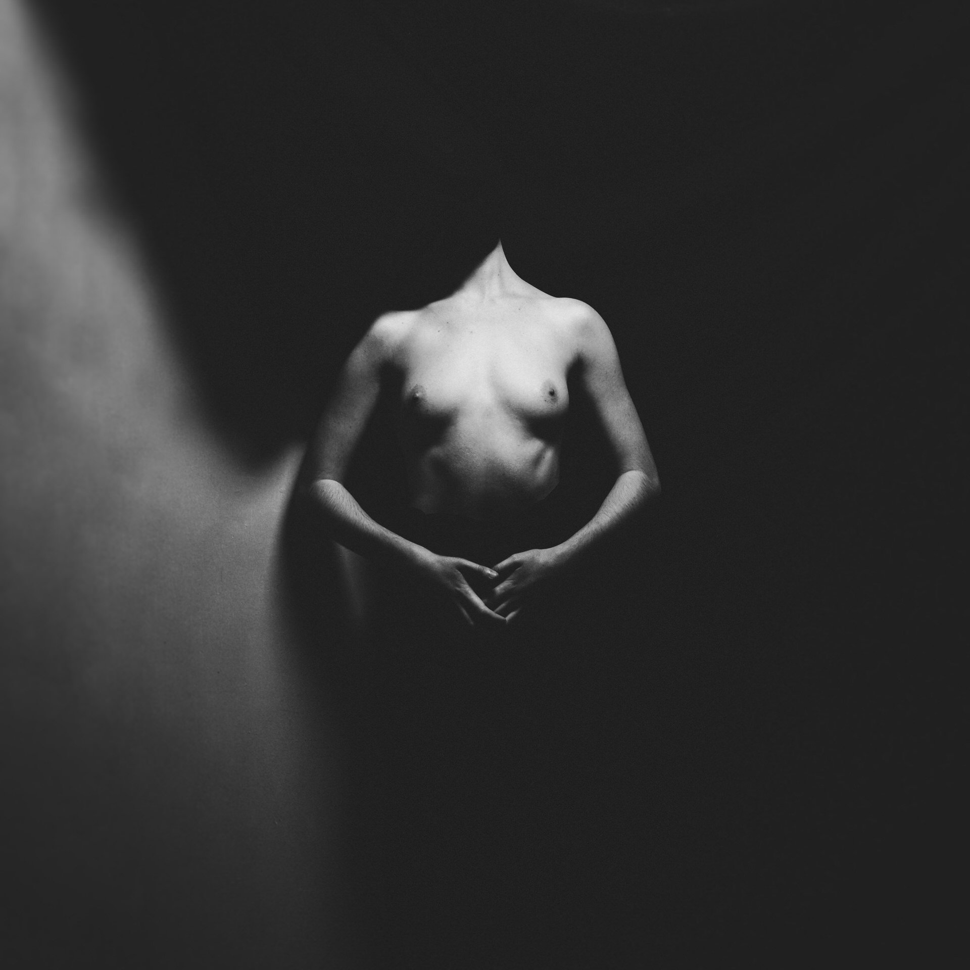 Photo en noir et blanc d'une personne nue sans tête, avec les bras croisés devant le ventre, dans un environnement sombre.