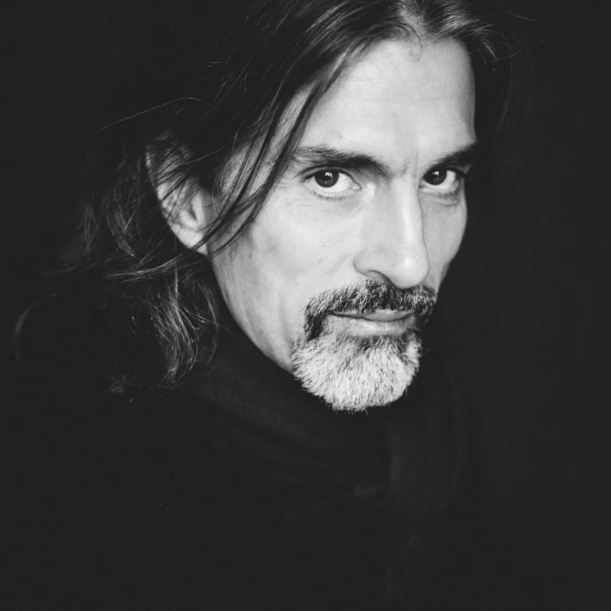 Portrait en noir et blanc d'un homme avec des cheveux longs, une barbe grise et un regard intense, regardant vers l'appareil photo.