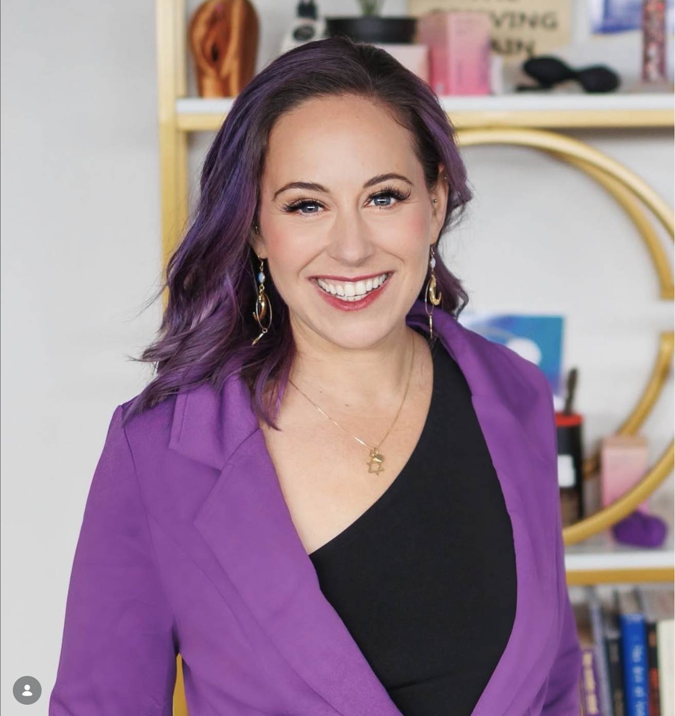 A woman with purple hair, wearing a purple blazer and black top, smiling, with jewelry including earrings and a necklace, in a room with a bookshelf and various decorative items.