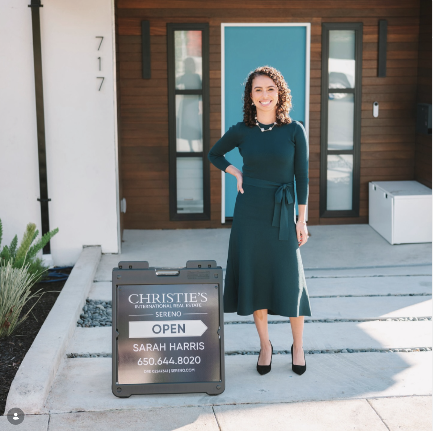A woman standing in front of a house with a sign indicating an open house for a property listed by Christie's International Real Estate. The woman is smiling, wearing a dark green dress and black heels.